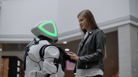 Smiling-girl-in-white-t-shirt-is-in-contact-with-humanoid.-Cyborg-helps-the-girl-answering-questions.-Useful-robot-assistant-in-the-office-and-shopping-center